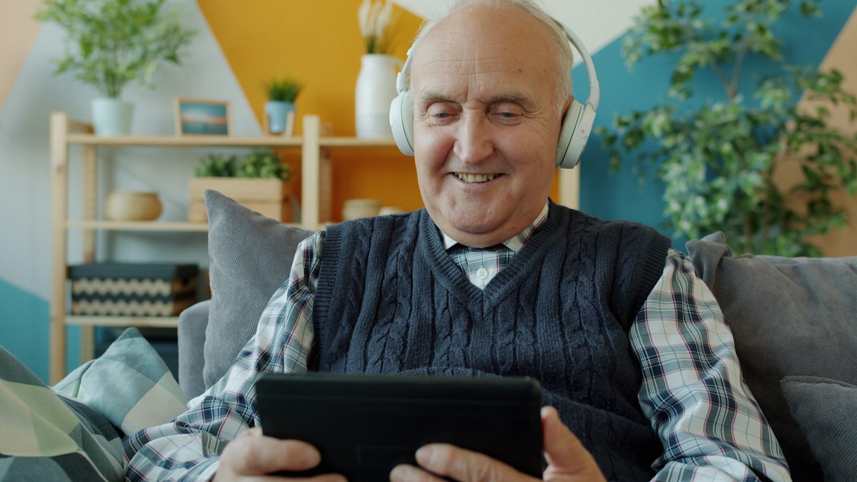 Homem idoso usa tablet e fones de ouvido em casa.