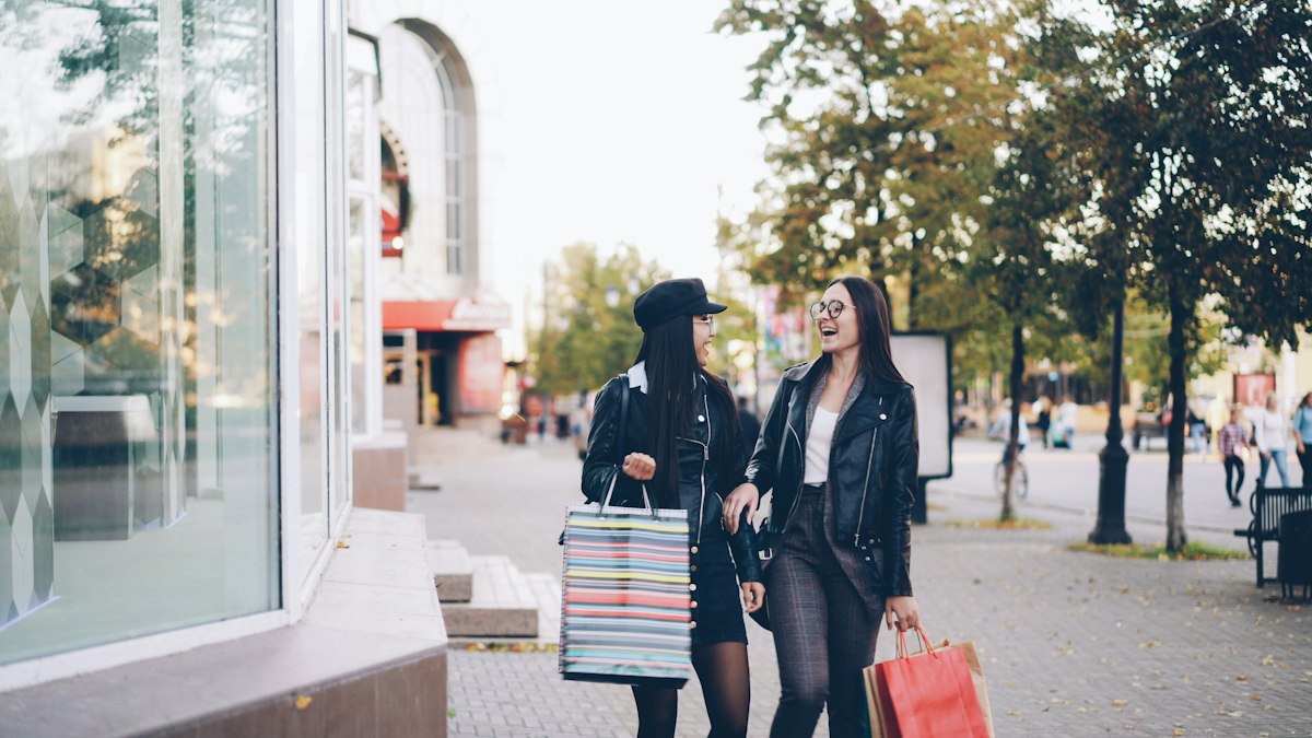 Duas mulheres caminham com sacolas junto a uma vitrine.